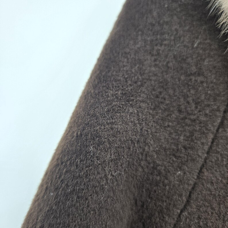 May include: Close-up of a dark brown coat with a textured surface. The coat appears to be made of a wool or wool-blend fabric. The edge of the coat is visible, with a light-colored fur trim.