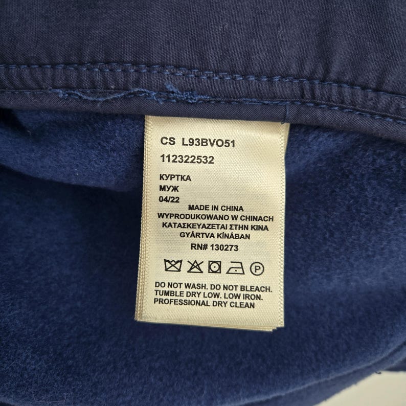 May include: A dark blue garment interior featuring a cream-colored care label with the text CS L93BVO51, 112322532, MADE IN CHINA, RN# 130273, and care instructions including DO NOT WASH, DO NOT BLEACH, TUMBLE DRY LOW, LOW IRON, PROFESSIONAL DRY CLEAN.