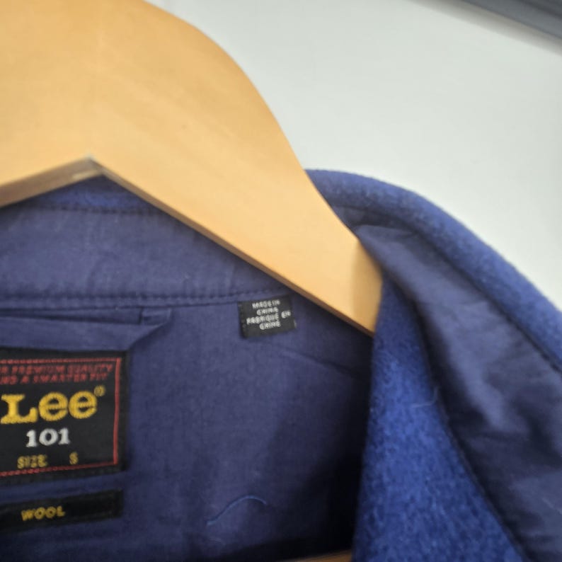 May include: Close-up of a navy blue wool jacket with a wooden hanger. The jacket features a Lee 101 label, size S, and a "WOOL" tag. A small tag reads "MADE IN CHINA".