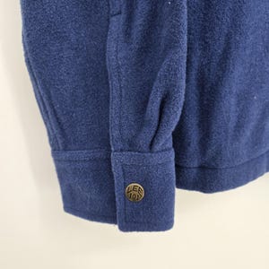 May include: A long-sleeved blue shirt made of a thick, felted fabric. The sleeve features a single-button cuff with a round, metallic brass-toned button embossed with the text Lee 101. The stitching is tonal, matching the blue fabric.