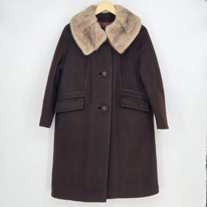 May include: A dark brown, knee-length coat with a light brown fur collar. The coat has two front pockets and is buttoned up. The coat is hanging on a wooden hanger against a white background.