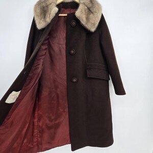 May include: A dark brown vintage coat with a fur collar and a burgundy lining. The coat has three buttons down the front, a pocket, and a tag on the inside. The coat is hanging on a wooden hanger.