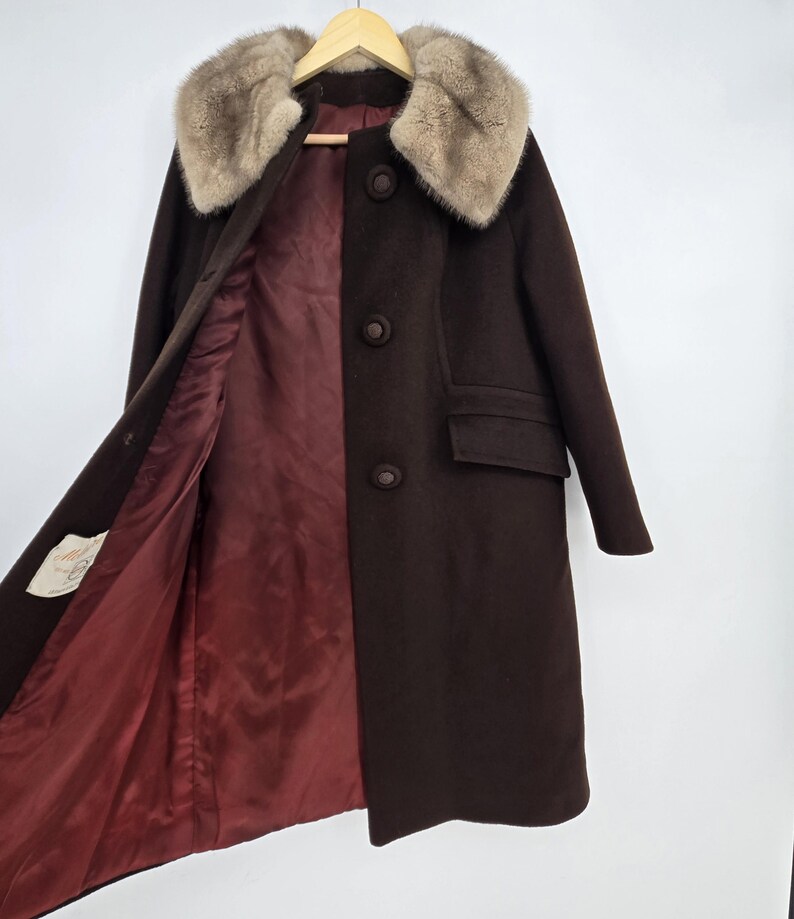 May include: A dark brown vintage coat with a fur collar and a burgundy lining. The coat has three buttons down the front, a pocket, and a tag on the inside. The coat is hanging on a wooden hanger.