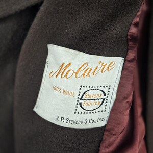 May include: Close-up of a brown wool coat with a label that reads "Molaire" in gold script. The label also states "100% Wool" and "Stevens Fabrics". The coat's interior lining is a deep red color.