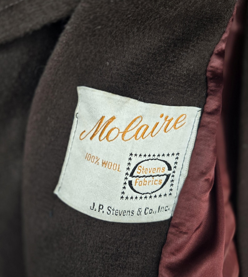 May include: Close-up of a brown wool coat with a label that reads "Molaire" in gold script. The label also states "100% Wool" and "Stevens Fabrics". The coat's interior lining is a deep red color.