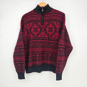 May include: A long-sleeve quarter-zip sweater featuring a black and red geometric pattern. The garment has a high collar, a metal zipper pull, and ribbed cuffs and hem. The intricate design includes repeating diamond and horizontal band motifs.