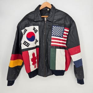 May include: A black leather jacket featuring a front design of multiple national flags including the South Korean, American, Canadian, and Italian flags. The sleeves display additional colour-blocked patterns in yellow, red, white, and blue.