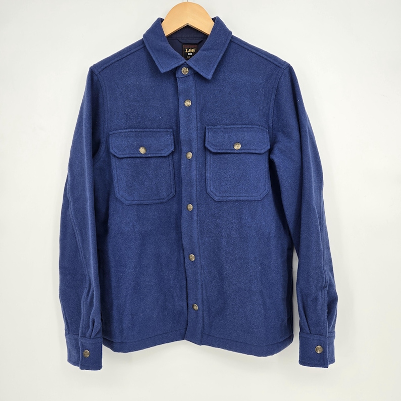 May include: A blue button-down shirt with two chest pockets and gold-colored buttons. The shirt is made of a soft, textured fabric and has a classic collar. The shirt is displayed on a wooden hanger.
