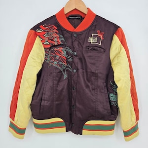 Puede incluir: Una chaqueta bomber de satén marrón oscuro con mangas acolchadas naranjas, detalles amarillos y un dobladillo acanalado con rayas verdes y rojas. El frente muestra bordados gráficos en rojo y verde y el texto Marithé François Girbaud en blanco.
