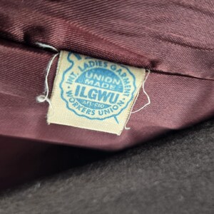 May include: Close-up of a vintage garment tag. The tag is light blue with the text "INT. LADIES GARMENT WORKERS UNION" and "UNION MADE ILGWU". The tag is attached to a dark burgundy fabric.