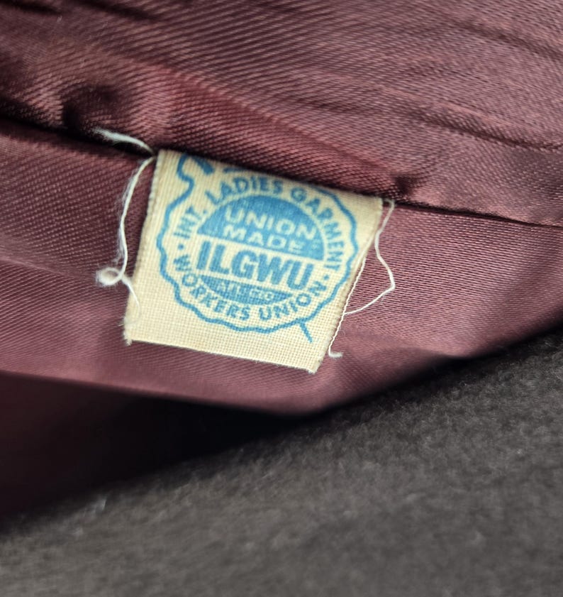 May include: Close-up of a vintage garment tag. The tag is light blue with the text "INT. LADIES GARMENT WORKERS UNION" and "UNION MADE ILGWU". The tag is attached to a dark burgundy fabric.
