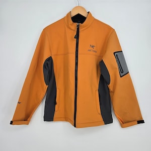 May include: An Arc'teryx softshell jacket in a vibrant mustard orange color with dark charcoal gray side panels. The jacket features a full-length black front zipper, a stand-up collar, a zippered chest pocket, and the Arc'teryx logo on the chest.