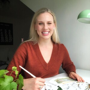 May include: A woman wearing a rust-colored sweater sits at a table and draws in a sketchbook with a white pencil. The sketchbook is open to a page with a drawing of a building. There are plants on the table.