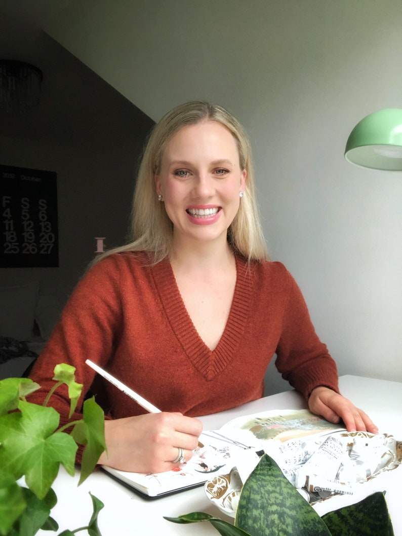 May include: A woman wearing a rust-colored sweater sits at a table and draws in a sketchbook with a white pencil. The sketchbook is open to a page with a drawing of a building. There are plants on the table.
