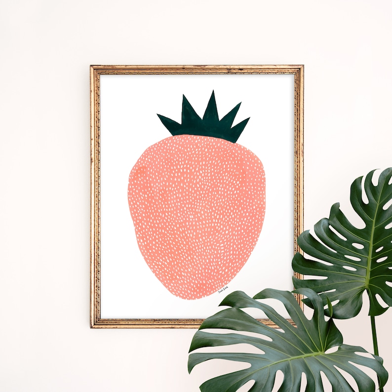 May include: A framed print of a stylized pink strawberry with white dots and a green leafy crown. The print is in a gold frame and is hanging on a white wall.