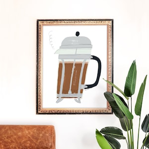 Op de afbeelding: Een ingelijste illustratie van een glazen French Press koffiezetapparaat met bruine koffie erin. De illustratie is in een gouden lijst en heeft een witte achtergrond. De tekst "coffee" is geschreven in een speels lettertype boven de koffiepot.