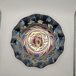 May include: Iridescent glass bowl with a ruffled edge. The bowl features a central design of a bird on a branch, surrounded by a wreath of leaves. The colours shift from blue to gold and purple.