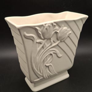 May include: A matte off-white ceramic vase with a rectangular, flared silhouette. The front features a raised, embossed relief of two tulips with stems and leaves, set against a background of diagonal lines. The vase has a solid, rectangular base.