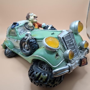 May include: A decorative figurine of a vintage-style light green car with white accents, featuring large black off-road tyres, a silver grille, and yellow headlights. A small figure with brown hair and a red shirt is seated in the driver's seat.