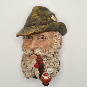 May include: A detailed wall sculpture of an elderly man with a long, curly white beard and mustache, wearing a green hat with a flower. He is smoking a pipe with a red cherry-like detail. The sculpture is painted with realistic skin tones.