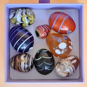 May include: A collection of eight small, oval-shaped glass beads with various patterns and colours, including striped, swirled, and spotted designs in shades of orange, black, white, yellow, and brown, arranged inside a square light purple box.