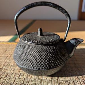 Könnte beinhalten: Eine schwarze Gusseisen-Teekanne mit einer strukturierten, genoppten Oberfläche. Die Teekanne hat einen gebogenen Griff und einen Ausguss. Der Deckel hat einen kleinen Knopf. Die Teekanne steht auf einer gewebten Matte.