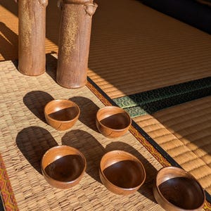 Puede incluir: Una colección de cerámica artesanal. Dos jarrones altos y cilíndricos con superficies marrones texturizadas y asas pequeñas. Seis cuencos pequeños y redondos de madera con interior oscuro. Los artículos están dispuestos sobre una estera tejida.