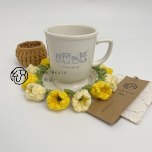 Può includere: Una tazza in ceramica bianca con un'illustrazione blu di quattro cagnolini e il testo "Our love is fur-real" poggia su un sottobicchiere rotondo all'uncinetto con fiori gialli e color crema e foglie verdi, accompagnata da un'etichetta marrone.