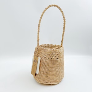 May include: A light tan woven straw bucket bag with a single braided handle and a scalloped top edge. A rectangular beige paper tag with black printed text and a circular logo is attached to the side of the bag with a thin brown string.