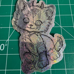 May include: A holographic sticker featuring a stylized cat with large eyes and tear-like drips. The cat has a distressed appearance with a split head and a dark, gradient color scheme. The sticker is on a green cutting mat.