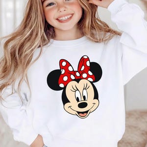 May include: White sweatshirt with a Minnie Mouse design. The graphic shows Minnie's face, a red bow with white spots, and black ears. The sweatshirt is a long-sleeved, casual top.