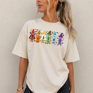 May include: A beige short-sleeve t-shirt featuring a colourful graphic print of six anthropomorphic crayons dressed in various Halloween costumes, including a vampire, pumpkin, ghost, witch, mummy, and wizard, surrounded by small bats and stars.