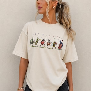 Op de afbeelding: Een crèmekleurig T-shirt met korte mouwen en een grafische illustratie van zes antropomorfe insecten die muziekinstrumenten bespelen, waaronder een kever, sprinkhaan, lieveheersbeestje, bij en krekel met zwevende muzieknoten.