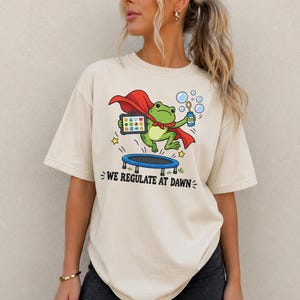 May include: A cream-colored t-shirt featuring a graphic of a green frog wearing a red cape, jumping on a blue trampoline while holding a tablet and a bubble wand. The text "WE REGULATE AT DAWN" is printed in black below the illustration.