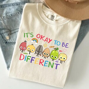 Peut inclure: Un t-shirt à manches courtes de couleur crème avec le texte IT'S OKAY TO BE DIFFERENT en lettres colorées. Le motif comprend des illustrations de dessins animés de diverses graines et fruits avec des visages, un petit arc-en-ciel, un soleil et un crayon.