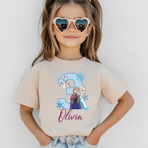 May include: A beige t-shirt featuring a blue number 3 with snowflakes and cartoon characters from the movie Frozen. The name "Olivia" is written in a cursive font below the number. The child is wearing heart-shaped sunglasses.