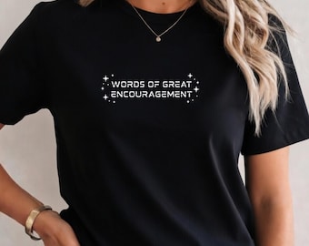 Può includere: Una t-shirt nera a maniche corte con il testo bianco "WORDS OF GREAT ENCOURAGEMENT" centrato sul petto. Il testo è affiancato da piccole icone a forma di stella a quattro punte su entrambi i lati, creando un design grafico sul tessuto nero.