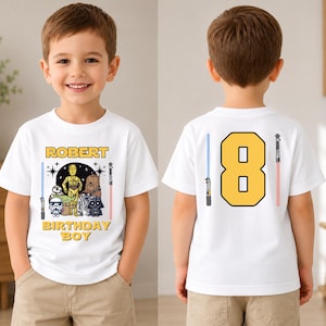 Pode incluir: Uma t-shirt branca com um gráfico de Star Wars na frente, o texto "ROBERT BIRTHDAY BOY" e várias personagens, incluindo C-3PO, Chewbacca e Darth Vader. As costas da t-shirt exibem um grande número 8 amarelo com sabres de luz.