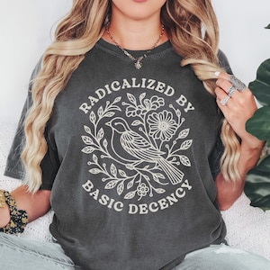 Pode incluir: T-shirt cinza escuro com o texto "RADICALIZED BY BASIC DECENCY" em um design circular. O design apresenta um pássaro, flores e folhas em um contorno branco. A camisa é um estilo confortável e casual.