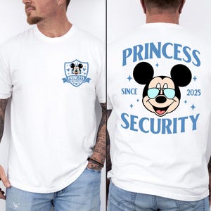 Pode incluir: Camiseta branca com um desenho do Mickey Mouse com óculos de sol e o texto "PRINCESS SECURITY" em azul. A frente tem um emblema menor com o mesmo design. A camiseta é de gola redonda e tem mangas curtas.