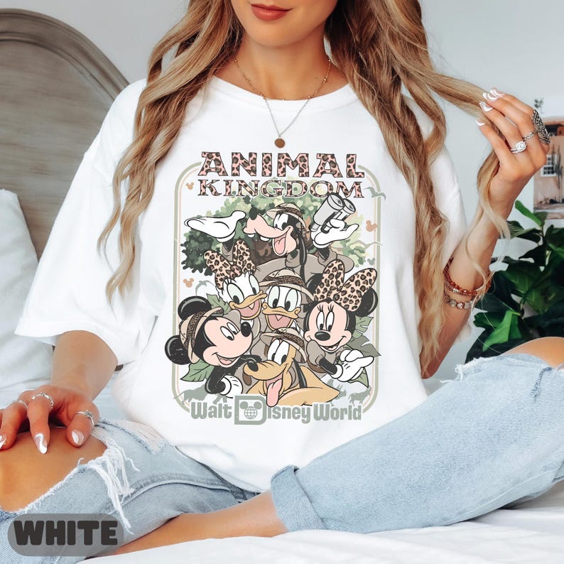 May include: White t-shirt with a graphic of Disney characters, including Mickey and Minnie Mouse, Donald and Daisy Duck, Goofy, and Pluto. The shirt has the text "ANIMAL KINGDOM" and "Walt Disney World" in a decorative frame.