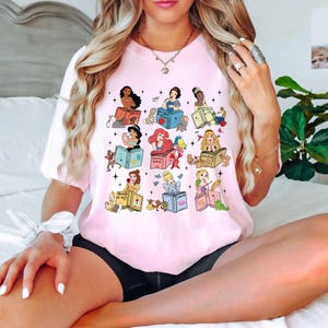 Peut inclure: T-shirt rose clair avec un motif color&eacute; de princesses Disney lisant des livres. Chaque princesse est repr&eacute;sent&eacute;e avec son propre livre, entour&eacute;e d'images associ&eacute;es. Le motif comprend des &eacute;toiles et des papillons.