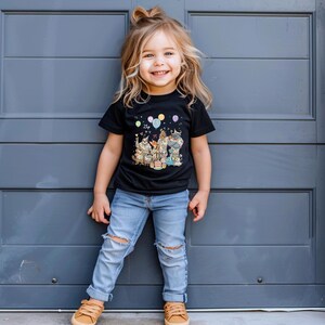 K&ouml;nnte beinhalten: Ein Kind tr&auml;gt ein schwarzes T-Shirt mit einem bunten Cartoon-Tier-Design, Luftballons und einer Geburtstagstorte. Das Kind tr&auml;gt auch hellblaue Jeans mit zerrissenen Knien und braunen Schuhen. Das Kind l&auml;chelt.