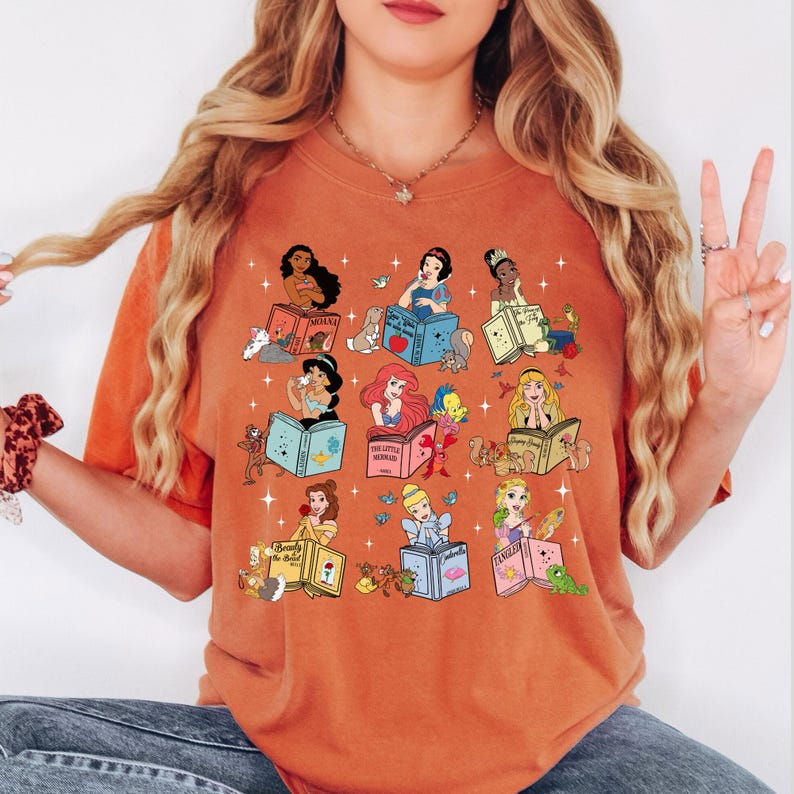 Peut inclure: T-shirt orange avec des illustrations de princesses Disney lisant des livres. Chaque princesse est repr&eacute;sent&eacute;e avec son nom et une image associ&eacute;e. Le t-shirt a une coupe d&eacute;contract&eacute;e et un col rond.