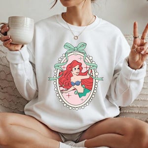 K&ouml;nnte beinhalten: Wei&szlig;es Sweatshirt mit einer Cartoon-Illustration einer Meerjungfrau in einem ovalen Rahmen, verziert mit einer mintgr&uuml;nen Schleife und einem Band. Die Meerjungfrau hat rotes Haar, einen blauen Muschel-BH und einen gr&uuml;nen Schwanz. Der Rahmen ist rosa mit einem gepunkteten Rand.