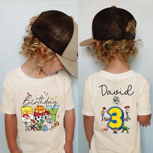 Pode incluir: Uma camiseta creme com o texto "Birthday BOY" e personagens de Toy Story. A parte de tr&aacute;s da camisa tem o nome "David" e um grande n&uacute;mero amarelo "3" com mais personagens de Toy Story. A crian&ccedil;a est&aacute; usando um bon&eacute; de caminhoneiro marrom e bege.