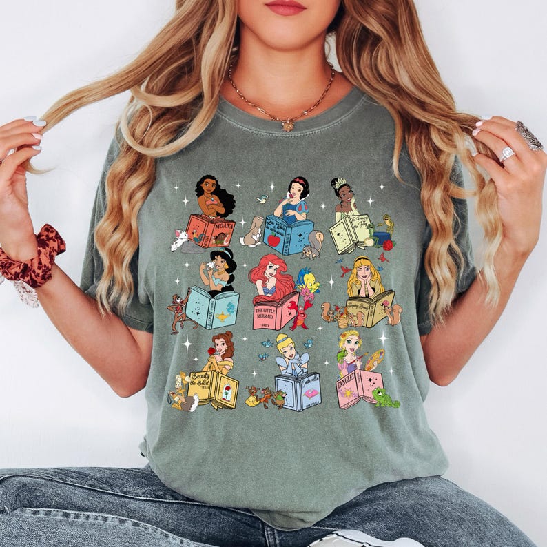Peut inclure: T-shirt vert sauge avec des illustrations de princesses Disney lisant des livres. Chaque princesse est repr&eacute;sent&eacute;e avec ses compagnons animaux et le titre de son livre. Le design comprend des personnages comme Ariel, Belle et Blanche-Neige. Le t-shirt est de style d&eacute;contract&eacute; et confortable.