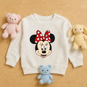 May include: Cream-coloured sweatshirt featuring a Minnie Mouse graphic. The graphic shows Minnie's face with a red bow with white polka dots. The sweatshirt is surrounded by a pink teddy bear, a yellow bunny, and a blue cat plush toy.