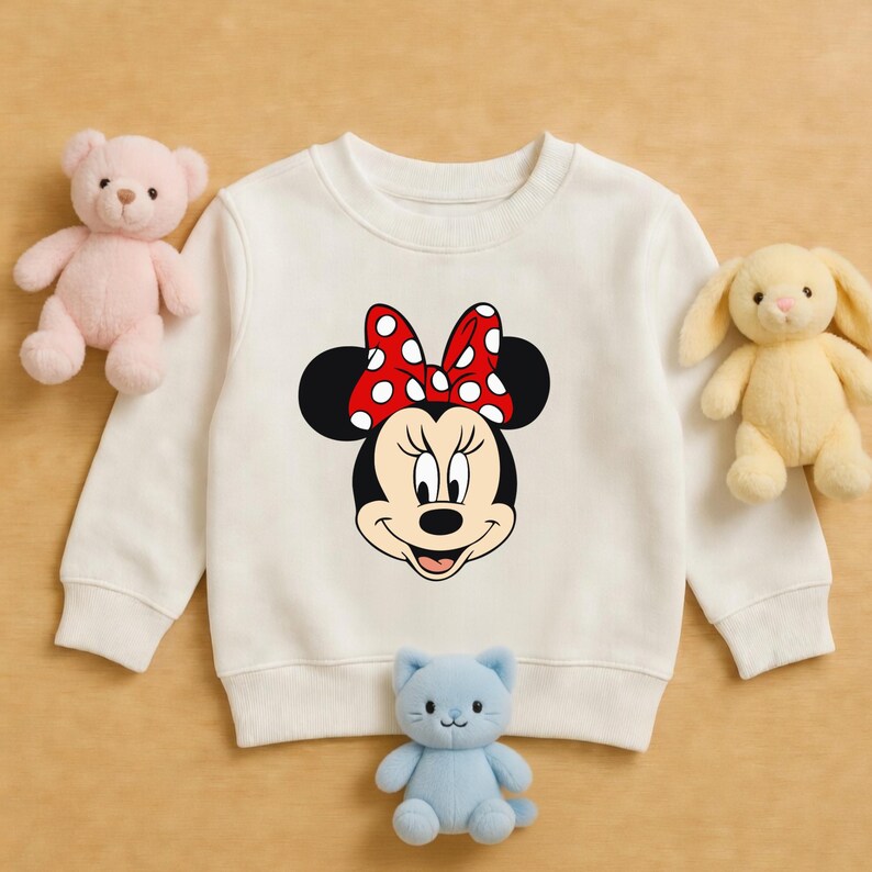 May include: Cream-coloured sweatshirt featuring a Minnie Mouse graphic. The graphic shows Minnie's face with a red bow with white polka dots. The sweatshirt is surrounded by a pink teddy bear, a yellow bunny, and a blue cat plush toy.