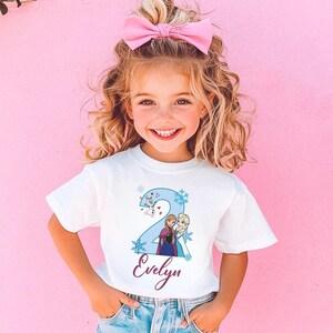 May include: A white t-shirt featuring a blue number 2 with cartoon characters and snowflakes. The name "Evelyn" is written in a cursive font below the number. The girl is wearing a pink bow.